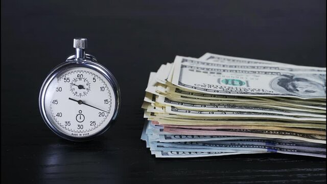 The stopwatch is lying on the table, and a hand is rapidly counting out dollar bills next to it, and the hand is ticking. The concept of sales, discounts and investments in stocks and businesses. time