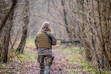 Naklejka premium Girl in military uniform with an airsoft gun walking on a dirt road in the forest