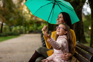 Mother and daughter sitting on bench in city park with umbrella.