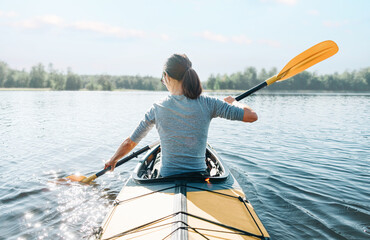 Young woman paddling in yellow color kayak on calm lake on sunny summer day, rear view.