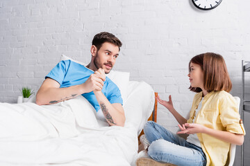 smiling girl gesturing while talking to father lying in bed in hospital