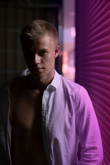 a young handsome guy stands in a poorly lit place near a shop window that gives off a lilac glow