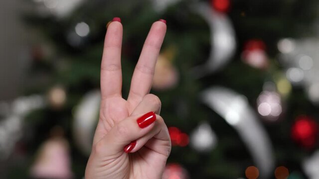 Woman raising two fingers up and showing peace or victory symbol or letter V. Female one hand holding two fingers up in sign language on holiday background. Fingernails with fresh red glossy manicure