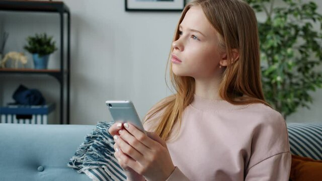 Serious female teenager is using smartphone texting touching screen sitting on couch in apartment alone concentrated on online communication. People and devices concept.