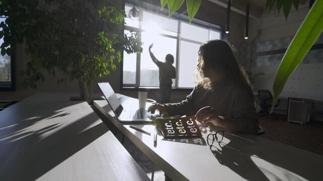 Businesswoman Asks Guy Silhouette And Talks Showing Laptop Display While Sitting At Long Office Table With Notebook And Pot Plants Under Back Sunlight