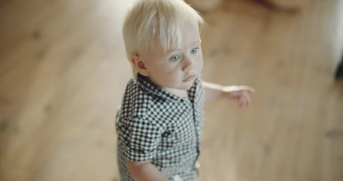 Serious baby boy walking down the room, looking thoughtfull and interested. Blond toddler with clear blue eyes. Wooden floor background.