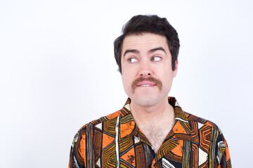 Amazed Young caucasian man wearing generic pattern printed shirt against white wall bitting lip and looking tricky to empty space.