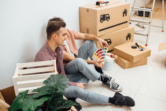 Young gay couple sitting and leaning against the wall of a new home they moved in, looking at color samples to choose the right color for their wall, while renovating apartment. Moving in a new home.