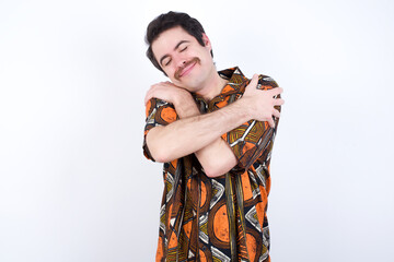 Young caucasian man wearing generic pattern printed shirt against yellow wall Hugging oneself happy...