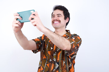 Young caucasian man wearing generic pattern printed shirt against yellow wall taking a selfie to...