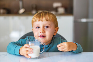 Adorable little boy drinking milk or yogurt and eating caramel having breakfast in the kitchen in the morning