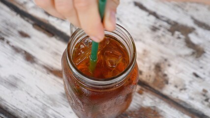 Top view of female hands stirring an ice tea in a jar with a plastic straw. Wooden table on a background 