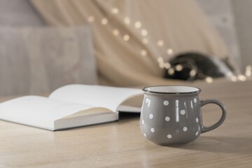 Cup of hot tea and book on the table, cozy home interior with sleeping cat on the background
