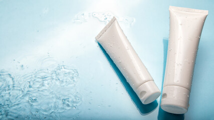 White cosmetic products in tubes on color blue background with water drops. Mock up skincare cosmetic packages on water surface. Natural moisturizing water balance cosmetics product. Long web banner
