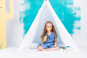 Portrait of a beautiful smiling girl with a paint roller. Little girl playing in a white wigwam on a background of colorful walls. Cheerful cute baby having fun in the nursery. baby's day. © Andriy Medvediuk