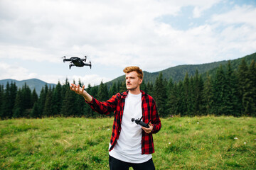 Fototapeta premium Handsome young man in a red shirt stands on a mountain meadow on a drone controls with a remote control on the background of a beautiful landscape. Guy flies a drone in the mountains.