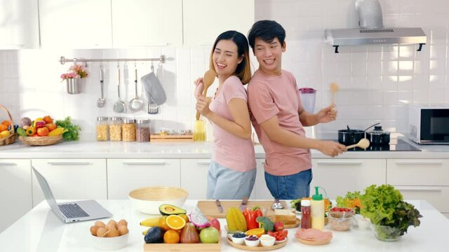 Happy Asian couple dancing laughing together preparing food at home, carefree joyful husband and wife having fun cooking healthy romantic dinner meal listen to music in modern kitchen. 4k high quality