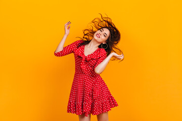 Stylish young girl in red short dress plays hair. Portrait of brunette in hoop earrings on yellow background