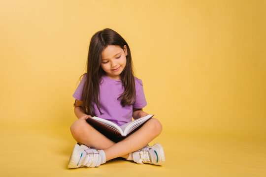 beautiful little girl in a purple t-shirt reads a book while sitting on a yellow background. Cute child learns lessons.