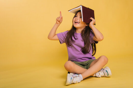 Beautiful Little Girl Holding A Book On Her Head While Sitting, Playing While Studying. Cute Baby Teaches Lessons On A Yellow Background. Photo In Studio