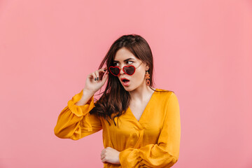 Shocked stylish lady takes off her heart-shaped glasses. Shot of brunette girl in bright blouse on isolated pink background