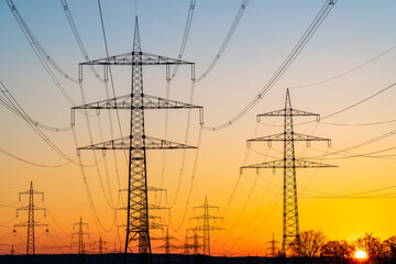 Fototapeta premium Electricity pylons and electric power transmission lines against vibrant orange sky at sunset. High Voltage towers provide power supply over a long distance for good distribution and infrastructure.
