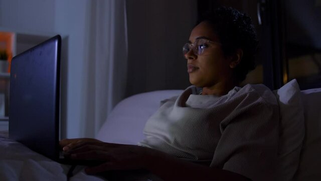 technology, internet, communication and people concept - young african american woman in glasses with laptop computer lying in bed at home at night