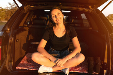 happy beautiful girl sitting in the trunk of car and enjoying sunset on the roadside. young woman...