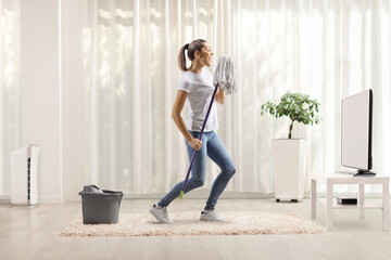 Happy young woman dancing and singing with a cleaning mop in front of tv