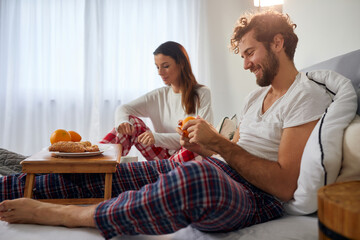 Obraz na płótnie Canvas A young guy with his girlfriend in the bed peeling off an orange for a breakfast. Love, relationship, together