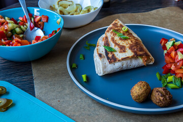 Fresh gluten free tortilla wrap on a plate stuffed with chickpea falafel, fresh vegetables chopped salad, fried onion rings, jalapenos, mayo and scallions. Healthy tasty meal preparation. Side view.