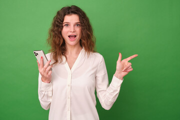 Beautiful girl in white clothes on a green background. The girl holds the phone in her hand, looks at the camera and points to an empty area with her index finger