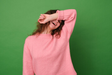 A cheerful carefree young girl laughs loudly holds her hand over her eyes tries to hide her face bursts into laughter isolated on a green background