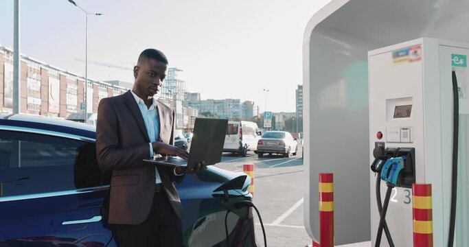 African American businessman holds a laptop in his hands and leans on a blue luxury electric car that is charging. Work While Charging The Car. the electric car is charged at an charging station