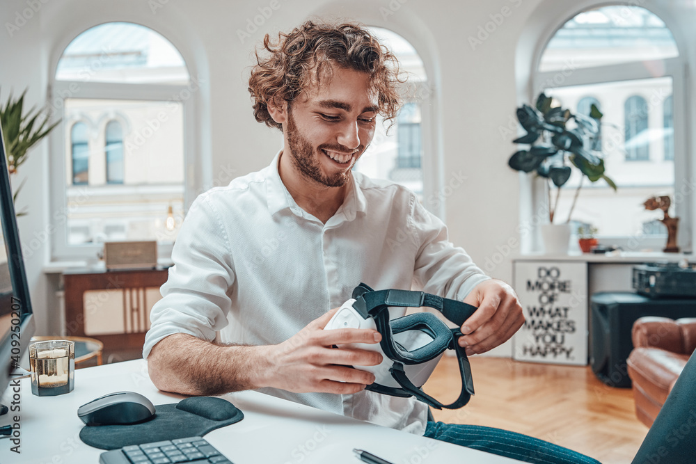 Poster portrait of a happy guy with curly hairs he sits at table near computer holding virtual reality head