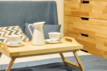 wooden tea table stands on the bed with cups. Bed with pillows and blue duvet and wooden chest of drawers