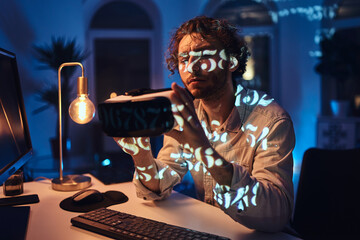 Portrait of a pensive caucasian guy in white shirt looking at virtual reality headset which he holds in dark room with lights.