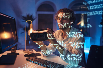 Atmospheric portrait of a astonished caucasian man with virtual reality headset sitting at table near computer in dark room with lights.