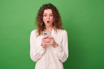 A girl on a green background in a white shirt holds a phone in her hands in surprise