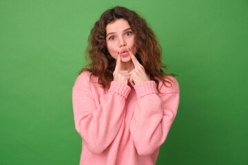 Beautiful young woman holds her index fingers near her folded lips makes a face at the camera wants to kiss someone wears a pink sweater isolated on a green background