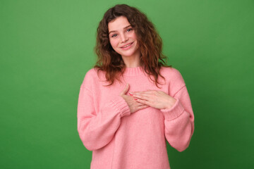 Happy woman presses her hands to her heart, expresses positive feelings, feels touched, makes a gesture of gratitude, wears a warm pink sweater, isolated on a green background