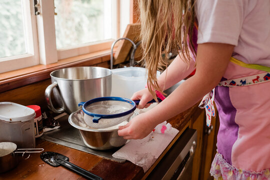 Girl wearing an apron and sifting flour into a bowl in the kitchen
