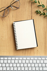 Blank paper notebook with keyboard, glasses, eucalyptus leaf on wood office desk table. Top view, flat lay.