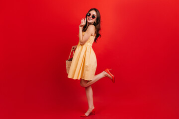 Snapshot full-length of pretty girl in short yellow dress on red background. Woman with dark wavy hair in sunglasses is smiling cute