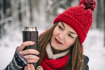 A beautiful woman in a red hat in a winter snowy forest looks at a thermos with hot tea.