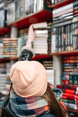 Obraz premium A beautiful woman standing with her back to the camera in a pink hat in a book store reaches for the top shelf to get a book.