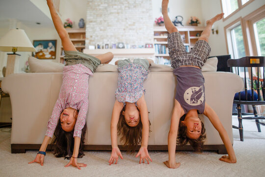 Three children lean against couch in living room doing handstands