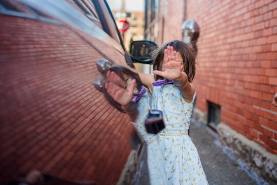 A little girl reflected in side of car holds hand up in front of face