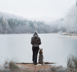 siberian husky dog sitting on the shore of a frozen lake with young female owner standing next to her in the mountains in winter