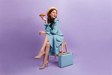 Full-length shot of young lady in elegant blue dress. Woman sits on stool next to suitcase and holds delicious croissant